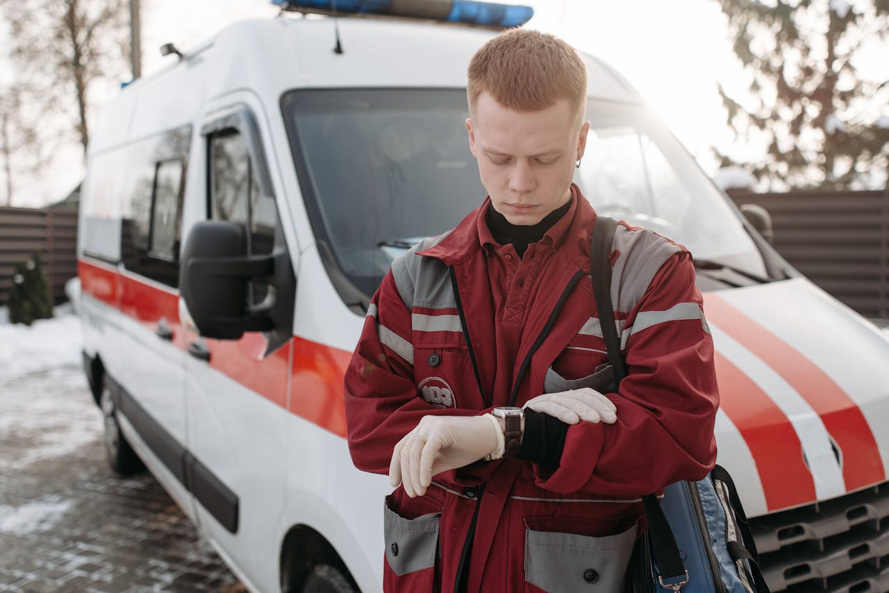A paramedic in uniform checks the time beside an ambulance outdoors on a sunny day.